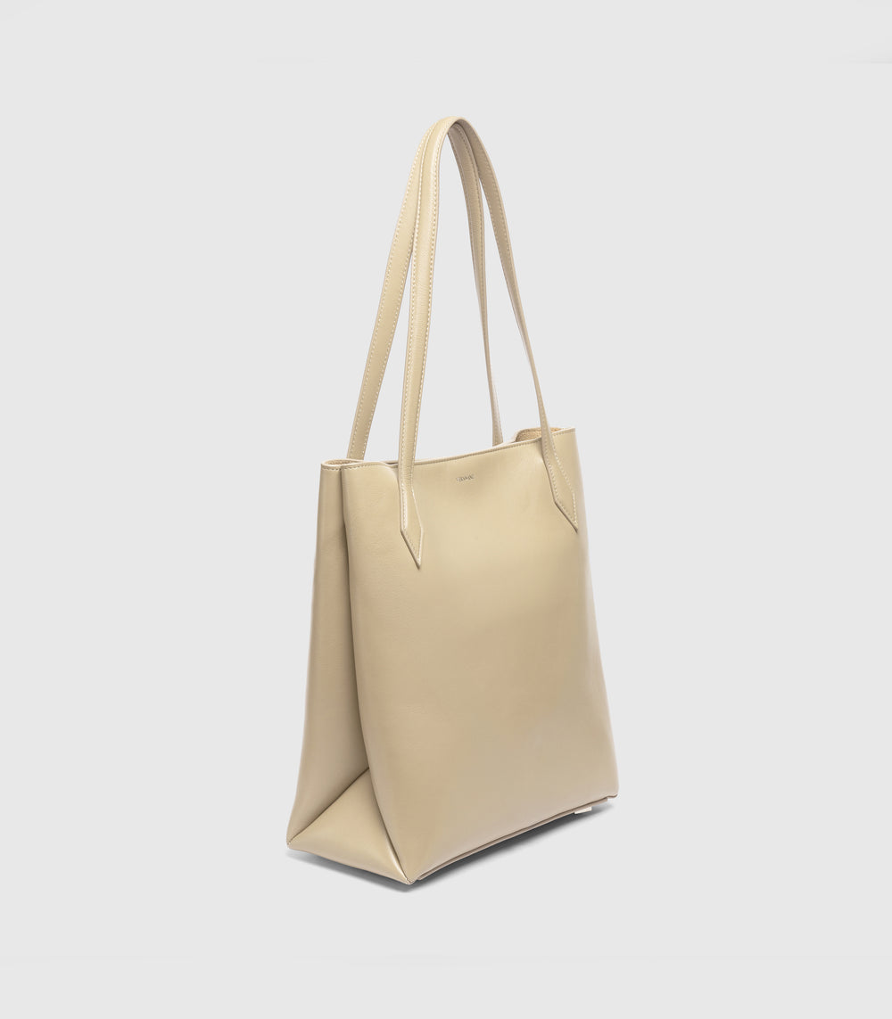 A light beige leather tote bag with long shoulder straps, shown from a three-quarters angle against a light gray background.
