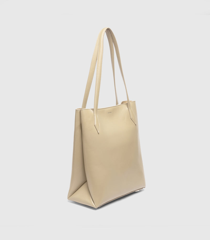 A light beige leather tote bag with long shoulder straps, shown from a three-quarters angle against a light gray background.