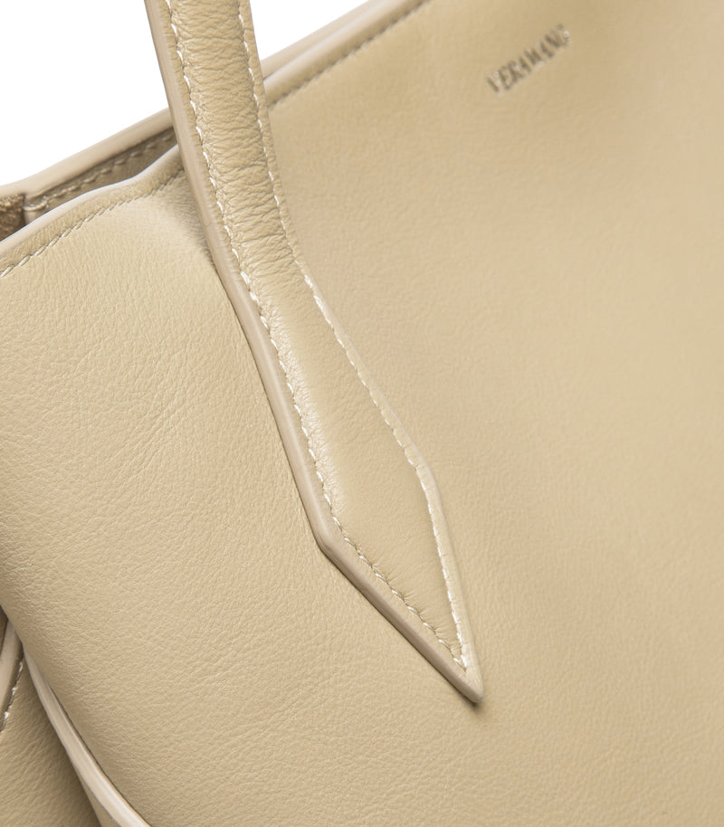 Close-up of a light beige leather handbag showing the pebbled texture, clean stitching, and a small debossed brand mark reading 'VERA WANG' on the upper right.