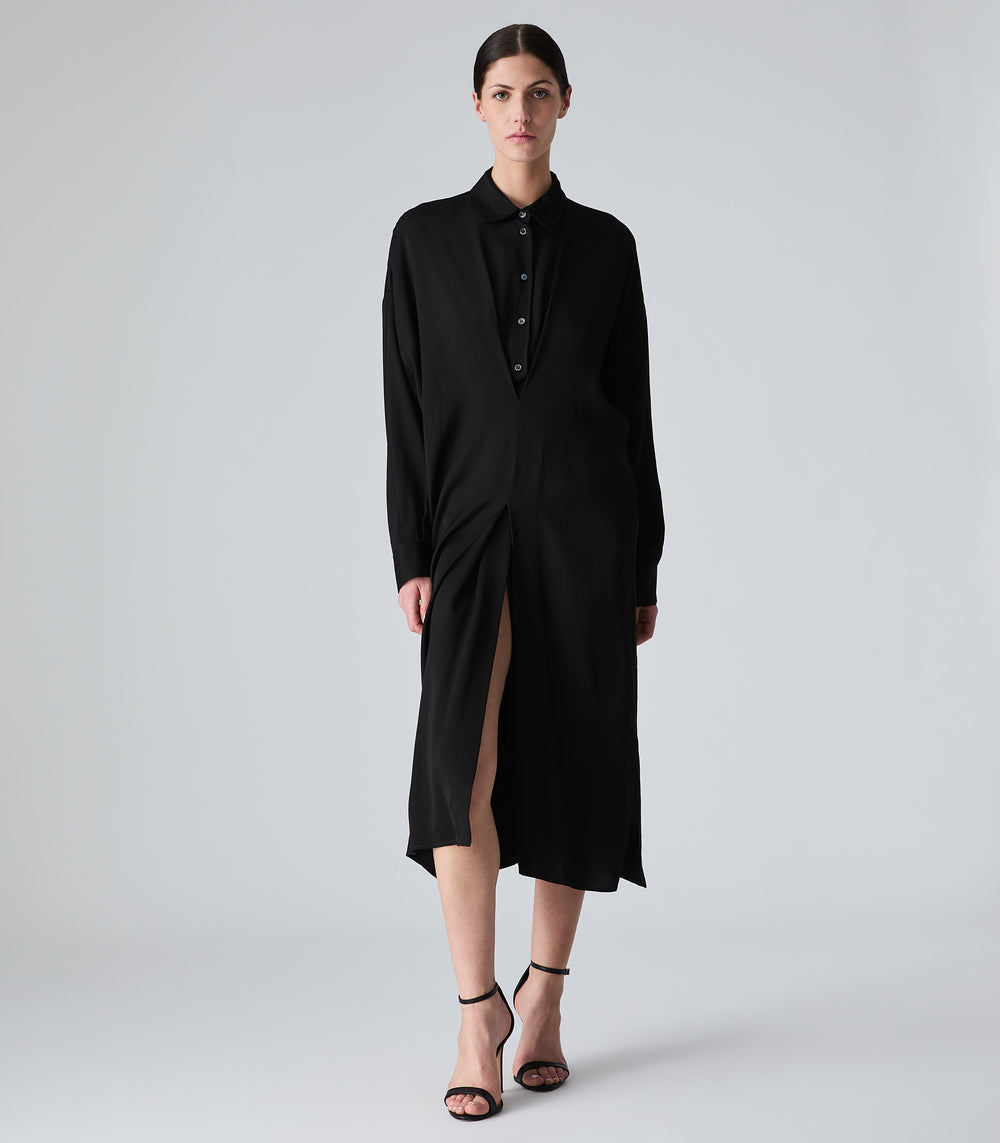 A model wearing a long black shirt dress with a high slit, standing against a light gray studio background.