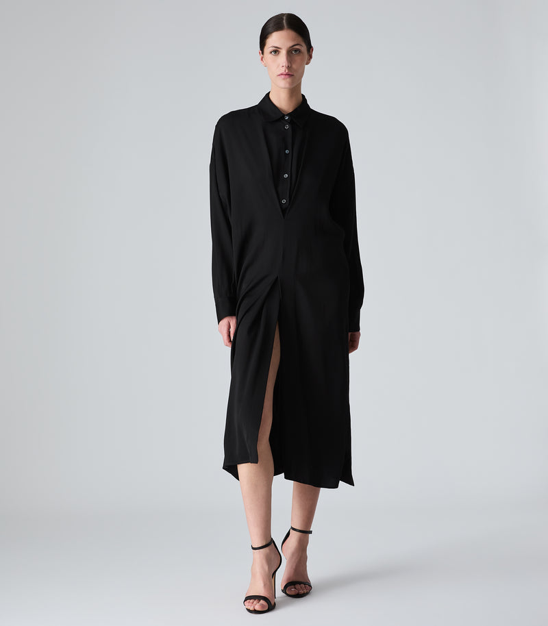A model wearing a long black shirt dress with a high slit, standing against a light gray studio background.