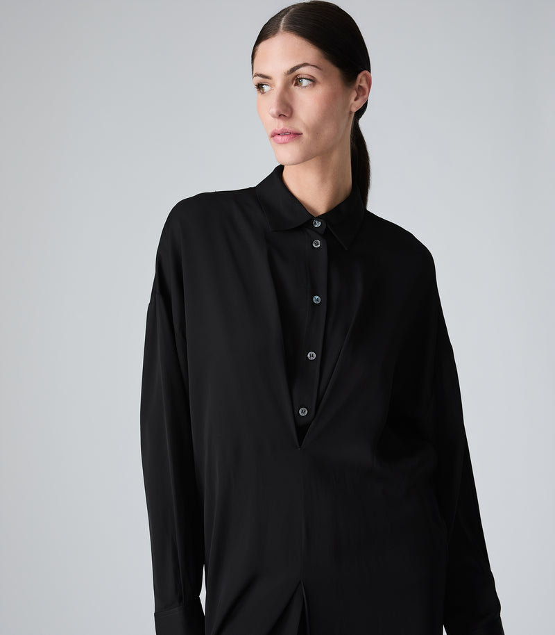 A woman wearing a black long-sleeve button-front shirt with a collar, photographed against a plain light gray studio background.