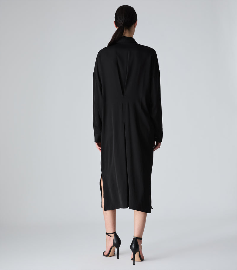 A model viewed from the back wearing a long black collared coat or dress with side slits and black high-heeled sandals, photographed against a light gray studio background.