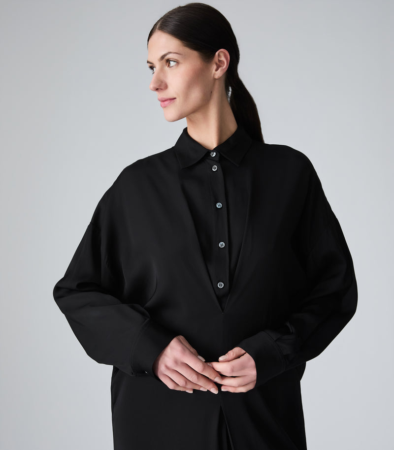 A woman wearing a black oversized button-down shirt with a collar and long sleeves, photographed against a plain light gray studio background.