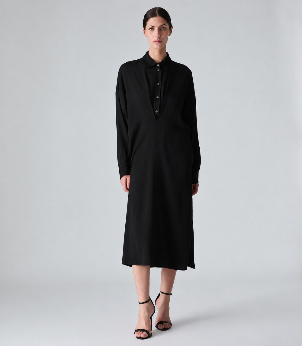 A model stands against a light gray studio background wearing a long-sleeved black midi shirt dress with a buttoned collar and black strappy high-heeled sandals.