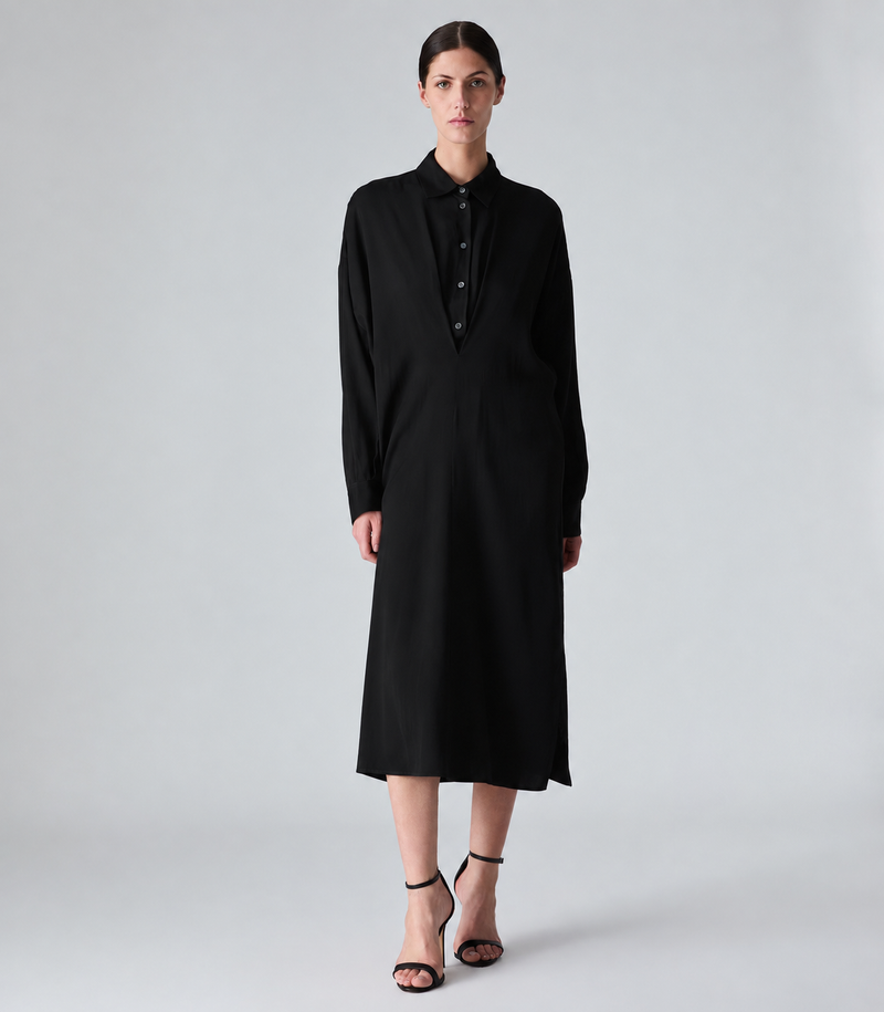 A model stands against a light gray studio background wearing a long-sleeved black midi shirt dress with a buttoned collar and black strappy high-heeled sandals.