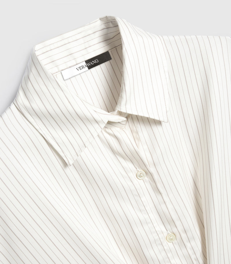 An angled, close-up product view of a shirt dress neckline, showcasing a classic pointed collar woven with fine vertical pinstripes in ivory and beige. The design features a hidden top button under a unique twisted fabric detail at the throat, a neat front placket with tonal four-hole buttons, and a visible split black-and-white inner neck 'VERA WANG' brand label against a light grey background.