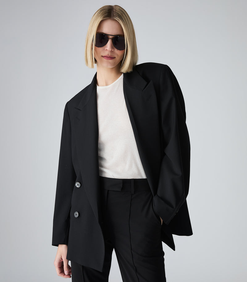 Woman wearing a black blazer and pants with sunglasses on a gray background