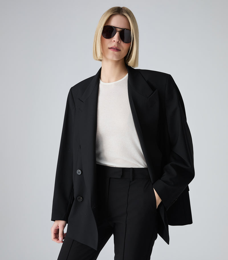Woman wearing a black blazer, white shirt, and black pants on a gray background