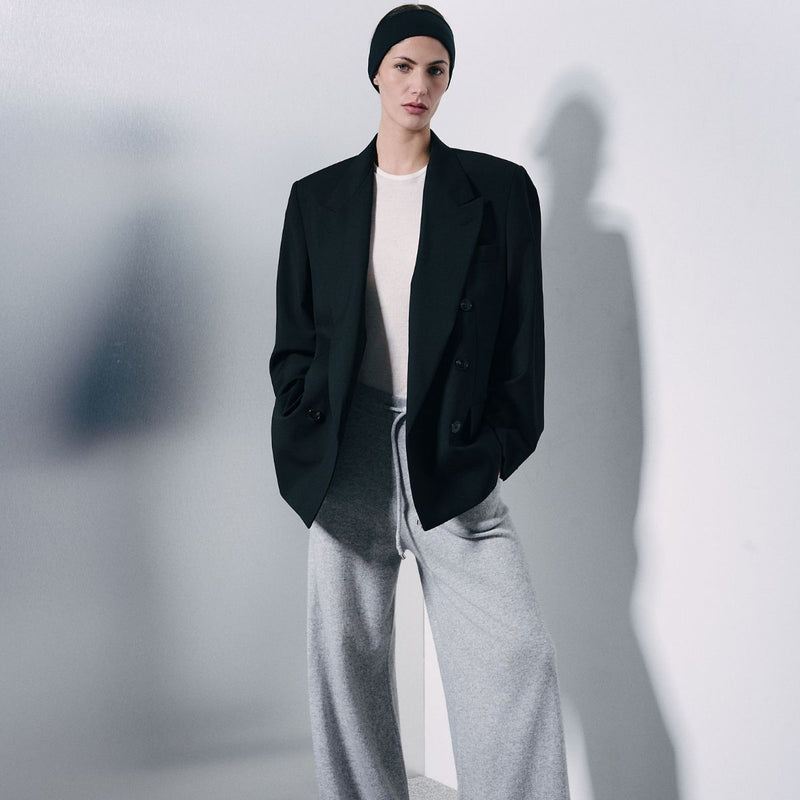A model wearing a black double-breasted blazer, white crewneck t-shirt, gray wide-leg sweatpants, and a black beanie, standing against a light gray wall with a soft shadow.
