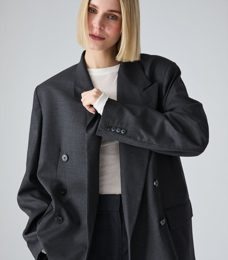 A blonde model wearing an oversized dark charcoal gray double-breasted blazer over a light beige top and dark trousers, photographed against a neutral light gray studio background.