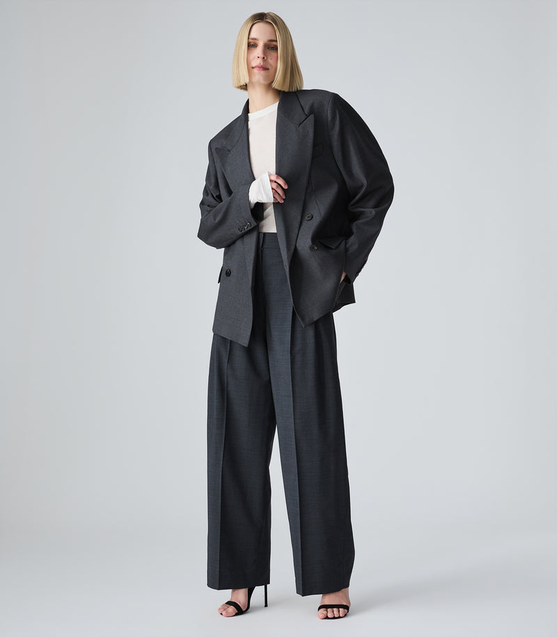 A model stands against a light gray studio background wearing a grey oversized blazer, matching wide-leg trousers, a white top, and black heeled sandals.