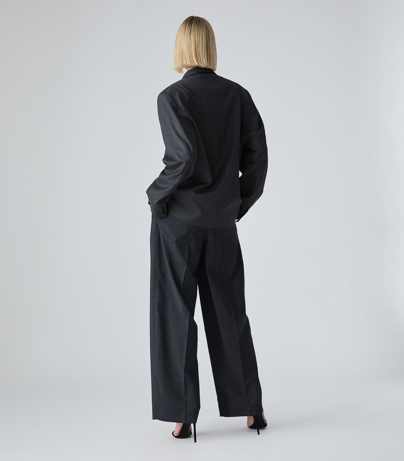 A model viewed from behind wearing a black long-sleeve collared shirt and matching wide-leg trousers, paired with black high heels, against a light gray studio background.