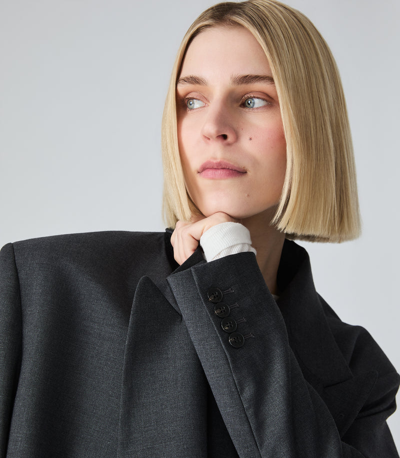 A blonde woman with a chin-length bob hairstyle wearing a dark charcoal gray oversized blazer over a white collared shirt, posing against a light gray studio background.