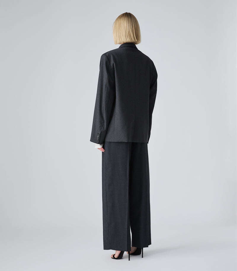 A model viewed from the back wearing a dark charcoal gray two-piece suit consisting of a long-sleeve blazer and wide-leg trousers, paired with black high heels on a light gray studio background.