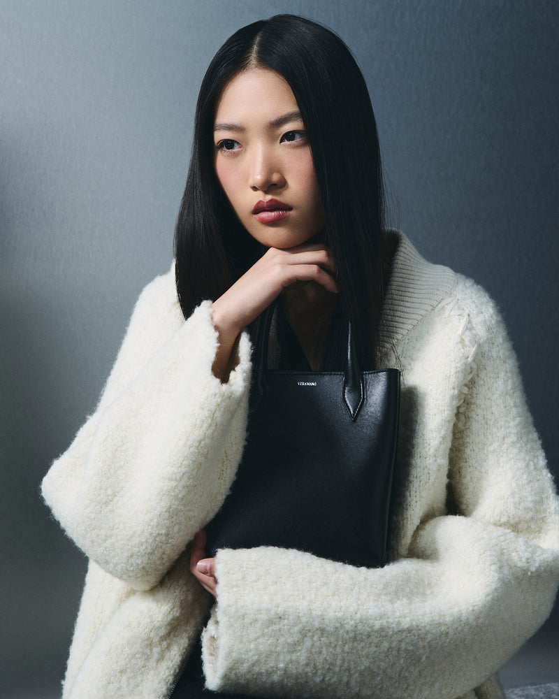 A model with long black hair is wearing a fluffy cream-colored knit cardigan and holding a structured black leather tote bag against a dark gray studio background.