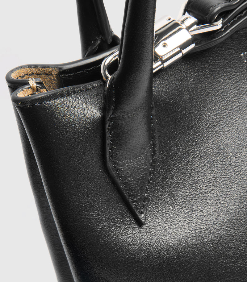 Close-up of a black leather handbag showing the pebbled texture, a black leather handle tab, silver-tone hardware, and a hint of tan interior lining against a light gray background.