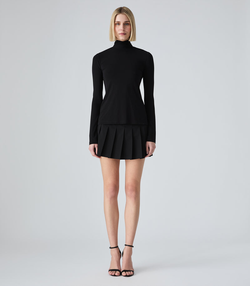 A model wearing a black long-sleeve mock-neck top, paired with a pleated black skirt and black strappy high-heels, against a light gray studio background.