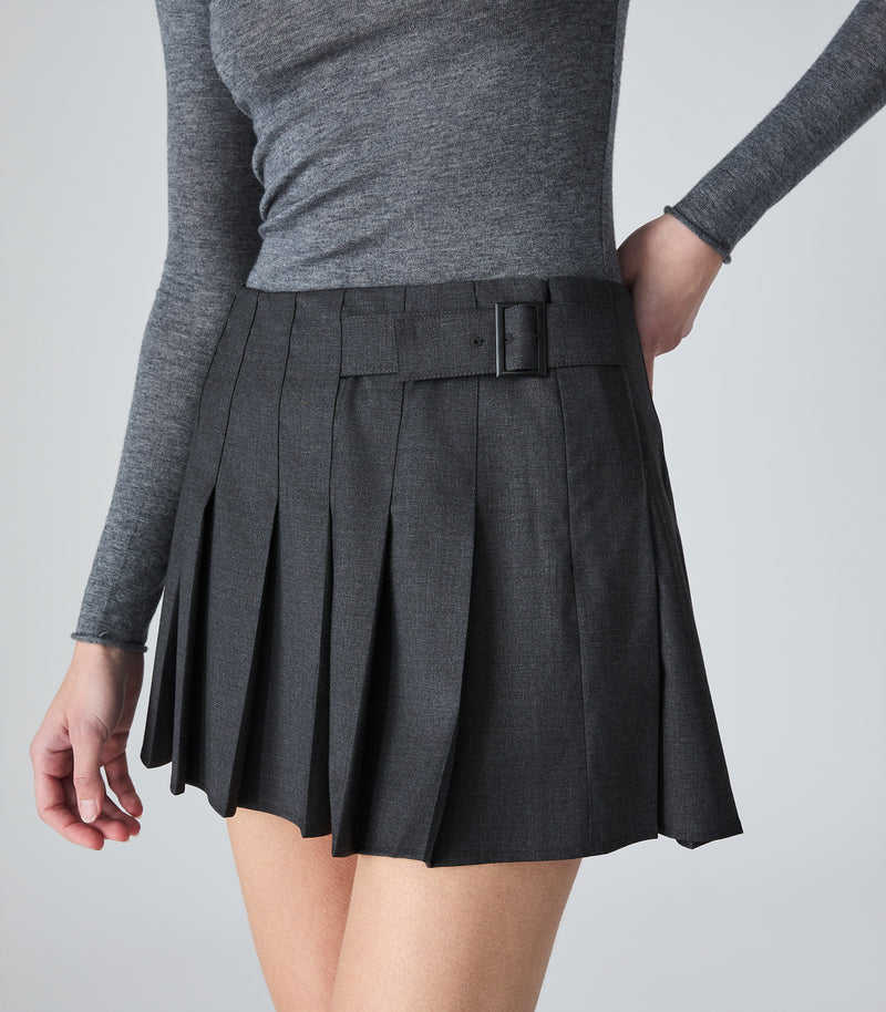 A model wearing a dark gray pleated mini skirt with a matching belt and buckle, paired with a gray long-sleeve top, shown from the waist down against a light gray studio background.