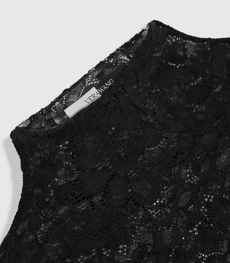 An angled, close-up product view of a high-neck top, highlighting the delicate transparency and openwork structure of the black floral lace fabric. The image focuses on the raw, scalloped edge of the mock neck collar and features a prominent split black-and-white inner neck 'VERA WANG' brand label positioned along the back neckline, against a light grey backdrop.