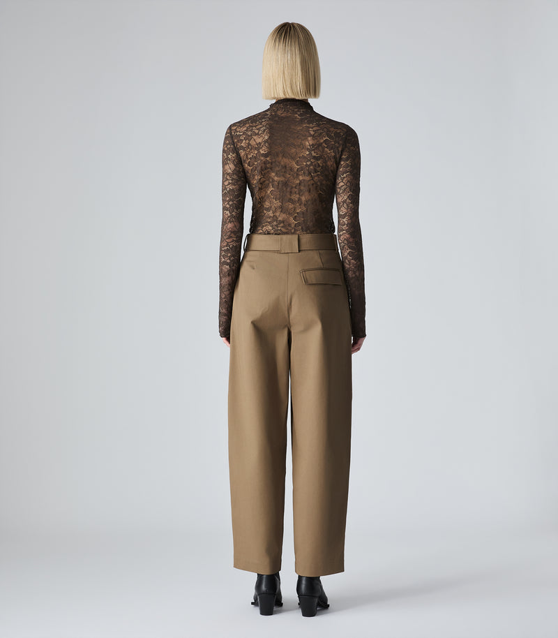 A model shown from the back wearing a sheer brown lace long-sleeve top and high-waisted tan wide-leg trousers, photographed against a light gray studio background.