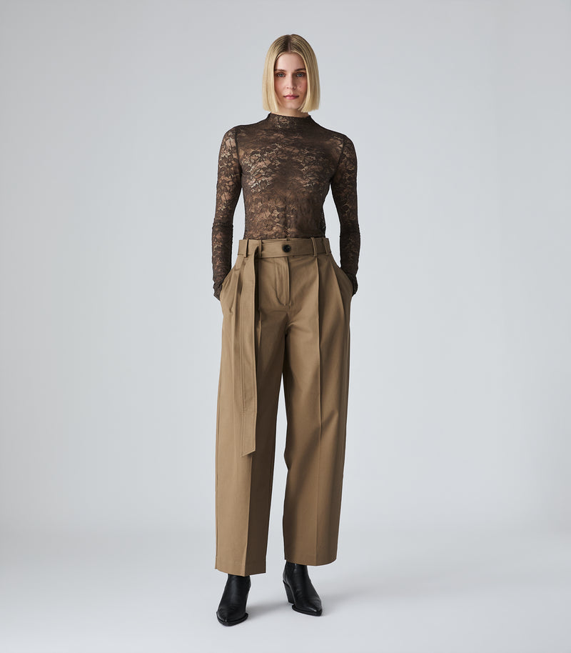 A model stands against a light gray studio background wearing a sheer black lace long-sleeve top paired with high-waisted, wide-leg tan trousers and black ankle boots.