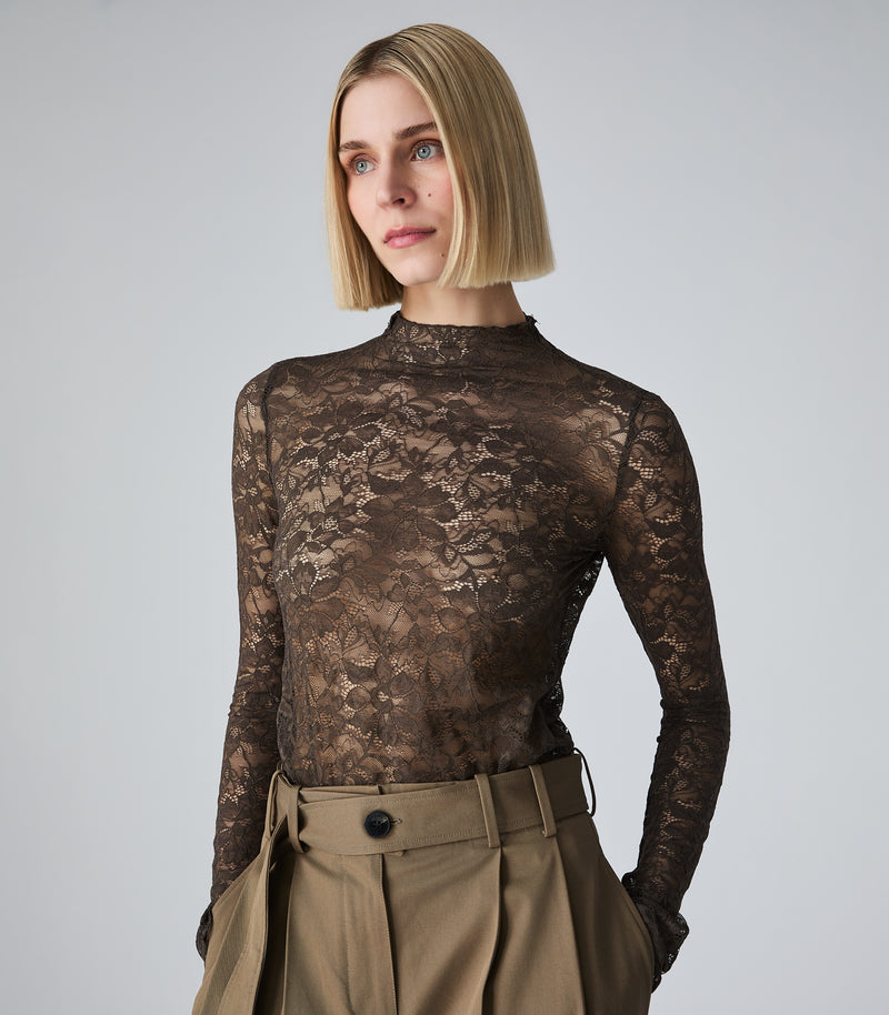A woman with a short blonde bob wearing a sheer brown floral lace mock-neck long-sleeve top and high-waisted khaki trousers, photographed against a neutral light gray studio background.