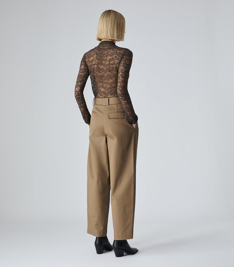 A model viewed from the back wearing a sheer brown lace long-sleeve top and tan wide-leg trousers, paired with black ankle boots, against a light gray studio background.