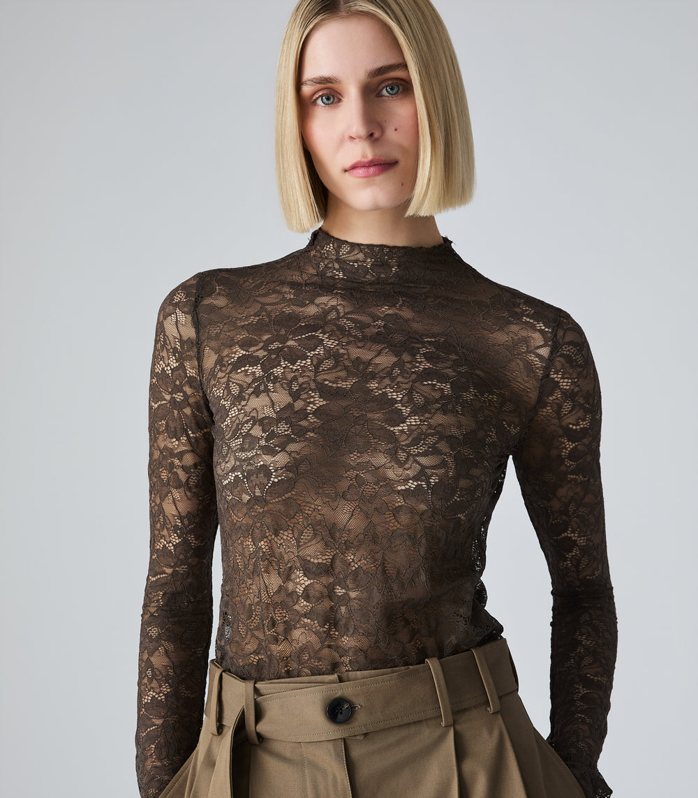 A woman with a blonde bob wearing a sheer brown lace mock-neck long-sleeve top and tan pleated trousers, photographed against a light gray studio background.