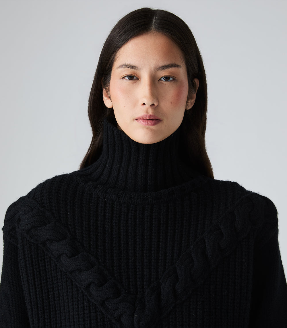 A close-up portrait of a model wearing a black ribbed turtleneck sweater with textured cable-knit details across the shoulders, set against a plain light gray background.