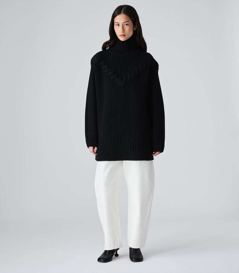 A model stands against a light gray studio background wearing a black ribbed turtleneck sweater with a wide draped collar detail, white wide-leg trousers, and black shoes.