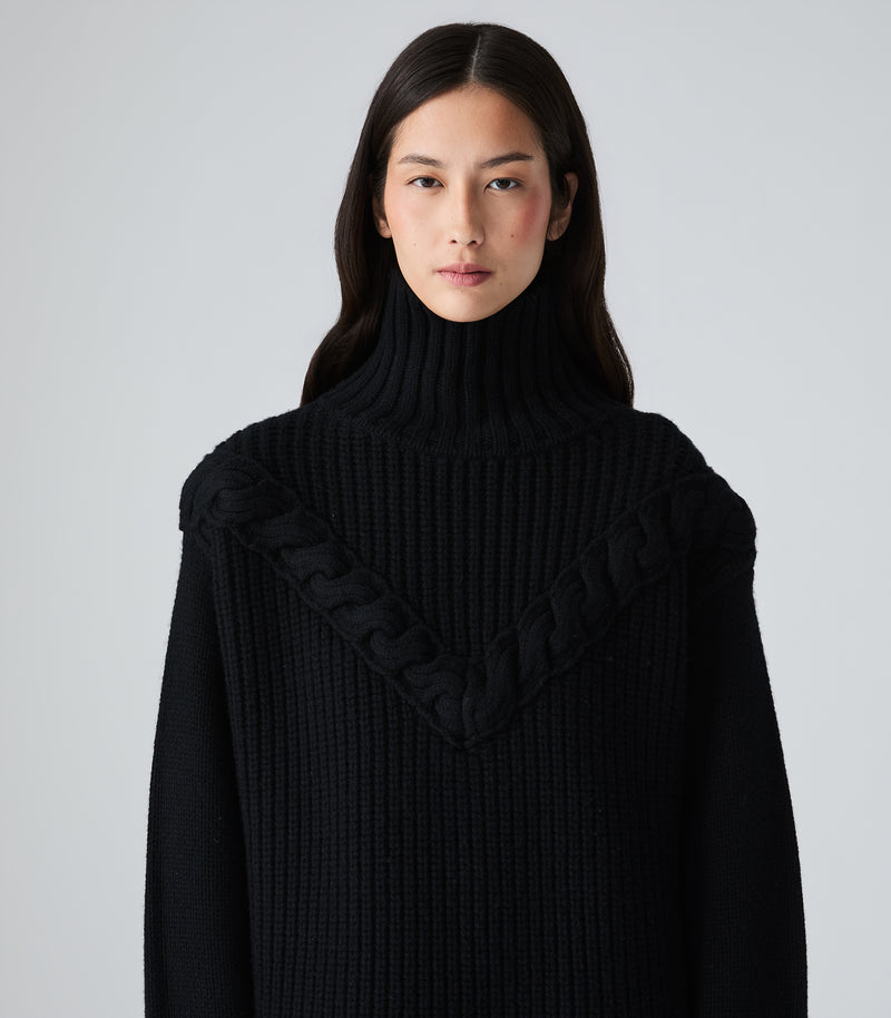 A model wearing a black ribbed knit turtleneck sweater with a braided V-neck detail, photographed against a neutral light gray background.