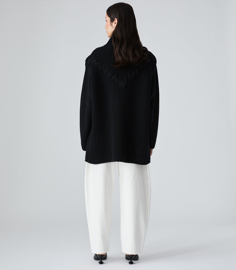 A model shown from the back wearing a black ribbed knit sweater with ruffle details on the shoulders and wide-leg white trousers, photographed against a light gray studio background.