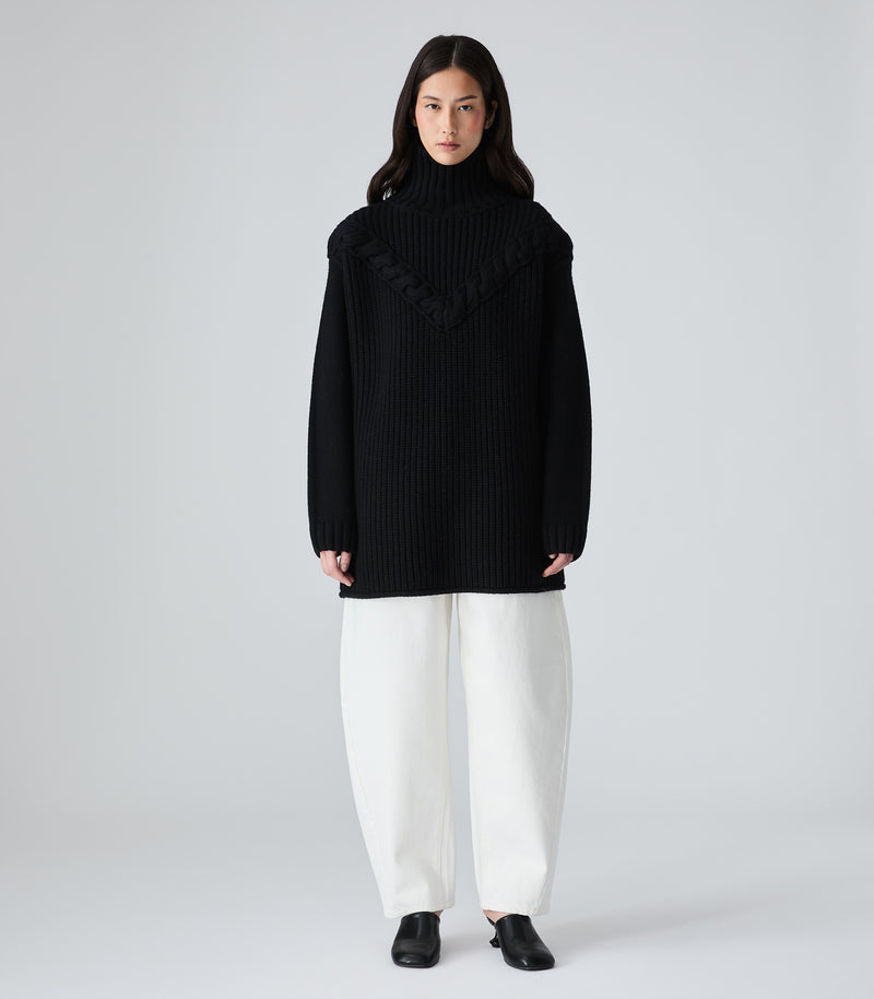 A model stands against a light gray studio background wearing a black ribbed turtleneck sweater with a wide draped collar detail, paired with wide-leg white trousers and black shoes.