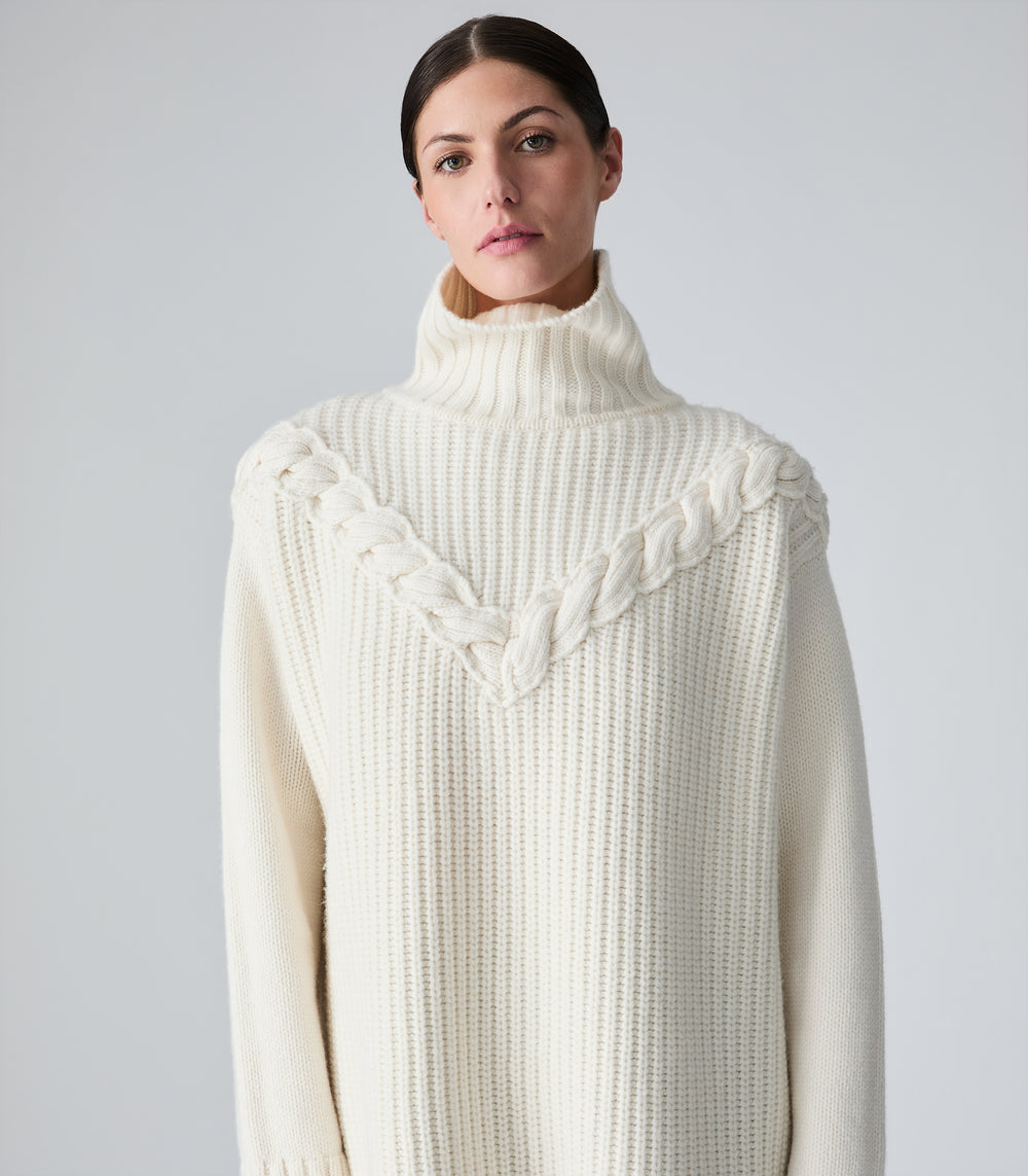 A woman wearing a cream-colored ribbed knit turtleneck sweater with a braided V-neck detail, photographed against a neutral light gray background.