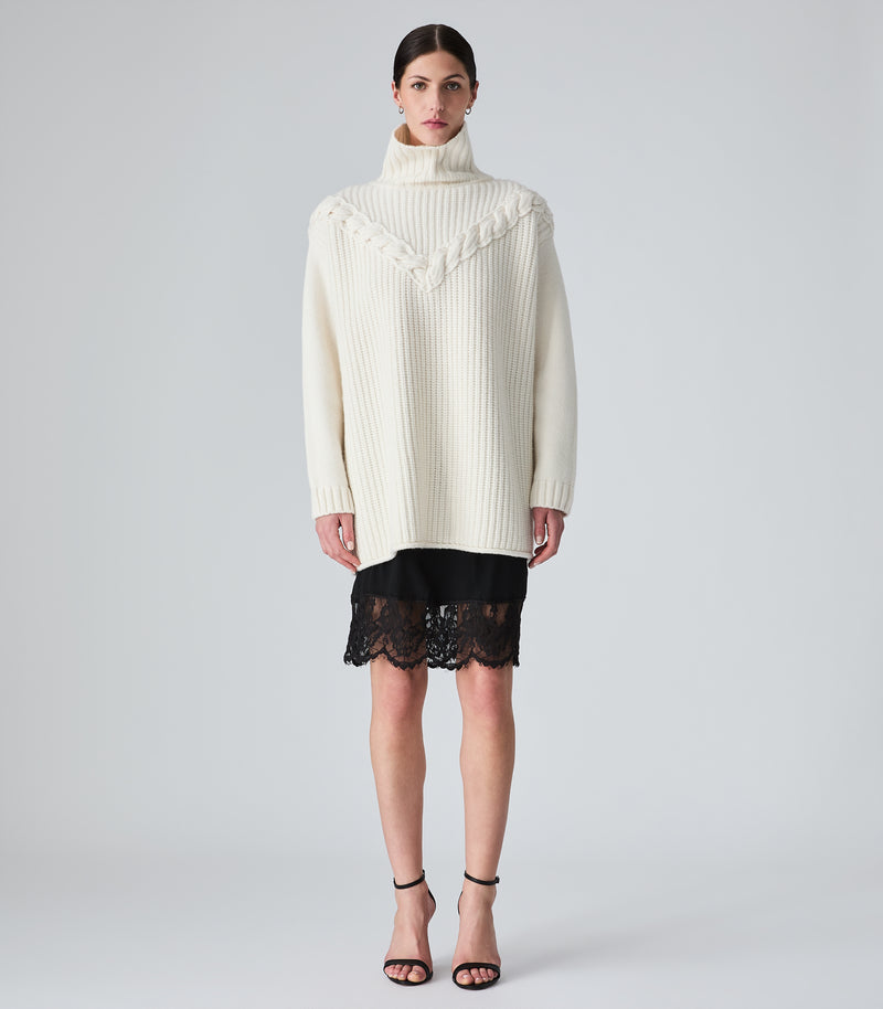 A model stands against a light gray studio background wearing a cream ribbed turtleneck sweater with decorative knit detailing across the chest, a short black lace skirt, and black strappy high-heeled sandals.