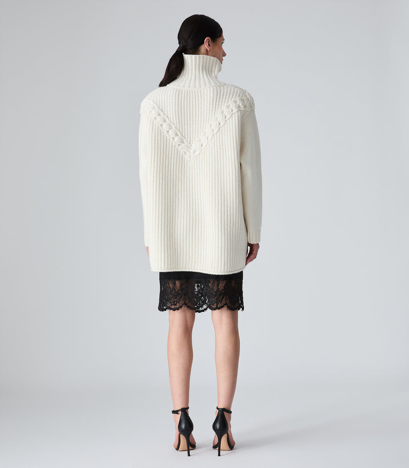 A model shown from the back wearing a cream ribbed turtleneck sweater with textured knit detailing across the shoulders, paired with a black lace mini skirt and black high-heeled sandals, against a light gray studio background.