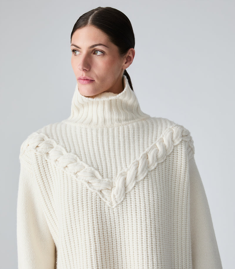 A woman wearing a cream-colored ribbed turtleneck sweater with a braided V-neck detail, photographed against a light gray studio background.