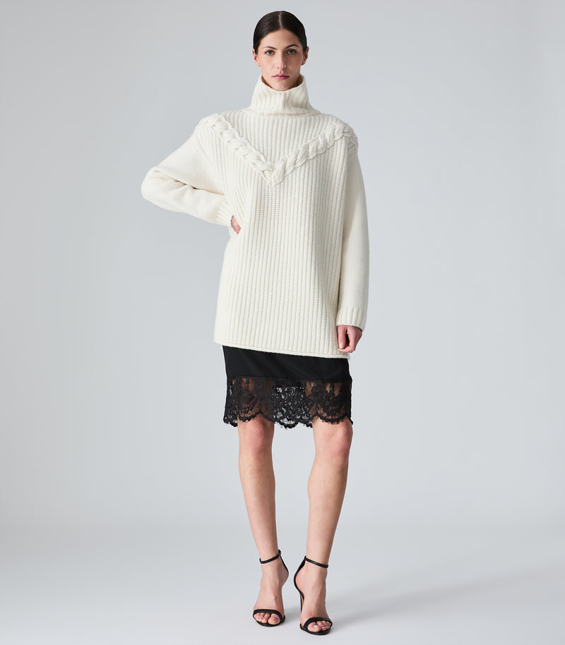 A model wearing a cream ribbed turtleneck sweater with decorative cable-knit detailing across the chest, paired with a black lace mini skirt and black strappy heels, against a light gray studio background.