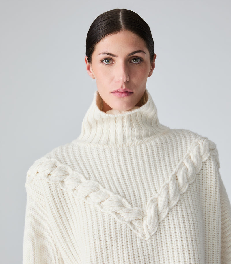 A woman wearing a cream-colored ribbed turtleneck sweater with braided shoulder details, photographed against a neutral light gray background.
