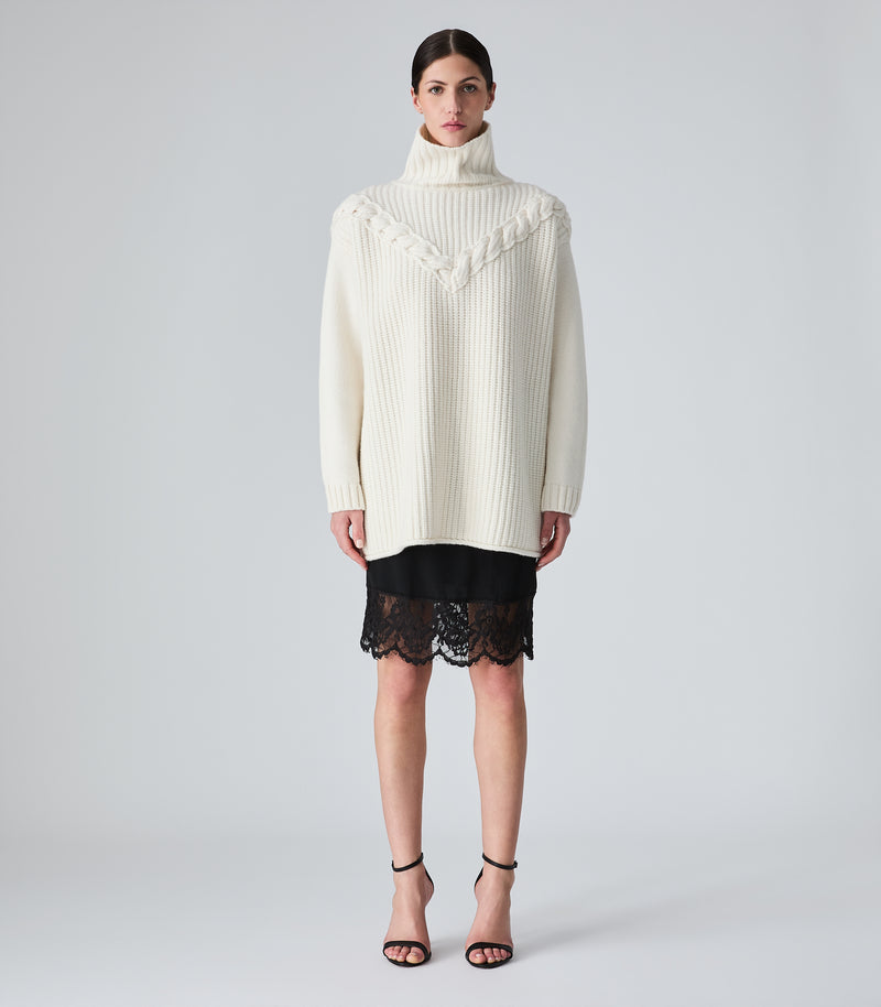 A model stands against a light gray studio background wearing an oversized cream ribbed turtleneck sweater with decorative yoke detailing, a short black skirt with a scalloped lace hem, and black strappy high-heel sandals.