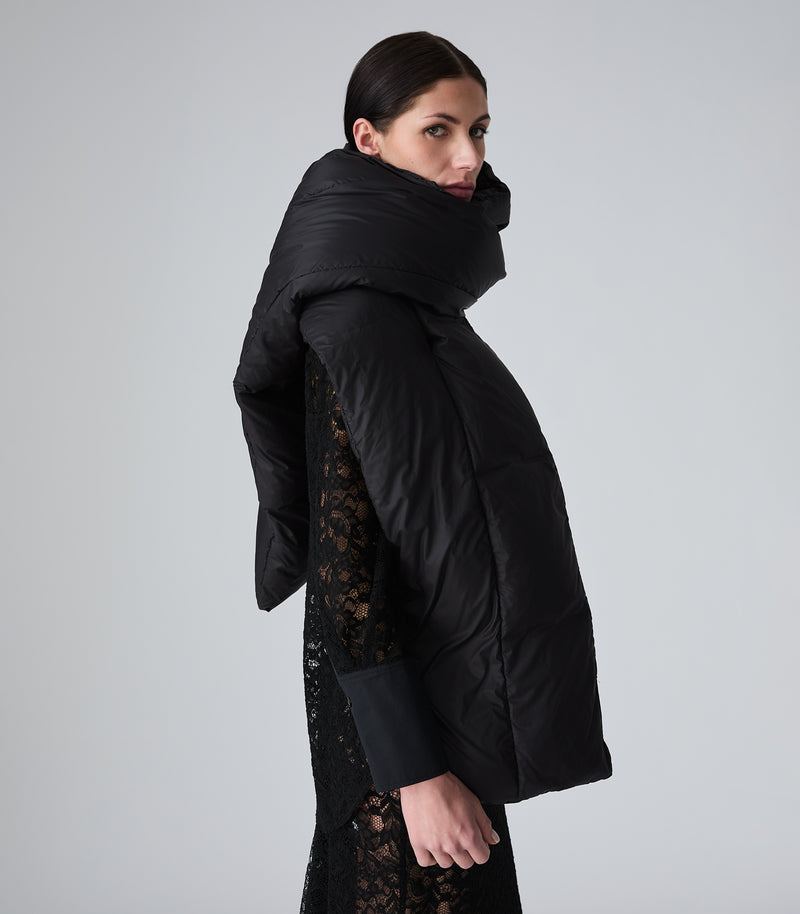 A model wearing a black puffer jacket with a high padded collar over a black lace dress, shown from a side-back angle against a light gray studio background.