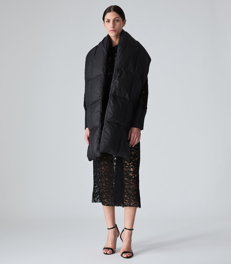 A model wearing a black puffer jacket with a high padded collar over a black lace dress, shown from a side-back angle against a light gray studio background.