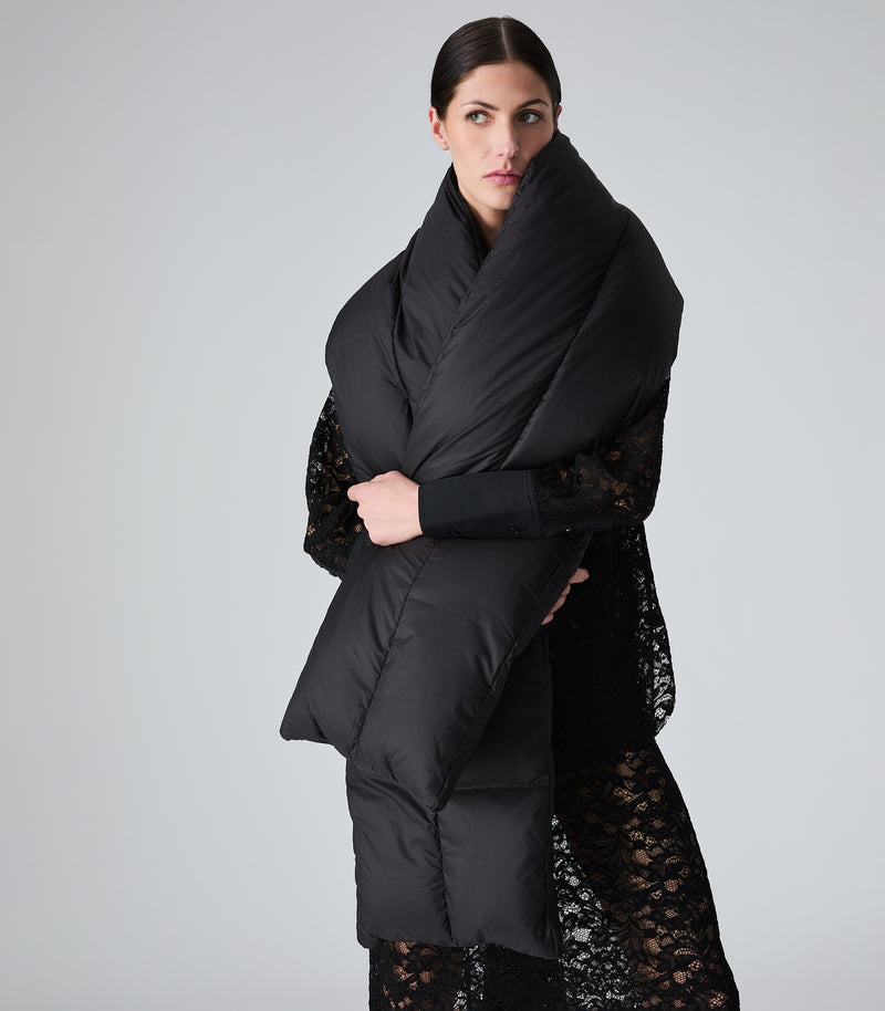 A model wearing a black puffer jacket with a high padded collar over a black lace dress, shown from a side-back angle against a light gray studio background.