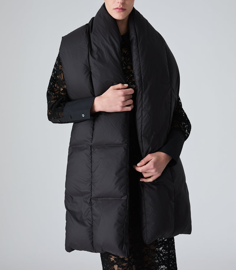A model wearing a long black puffer vest over a black lace top, shown from the chin down against a light gray studio background.