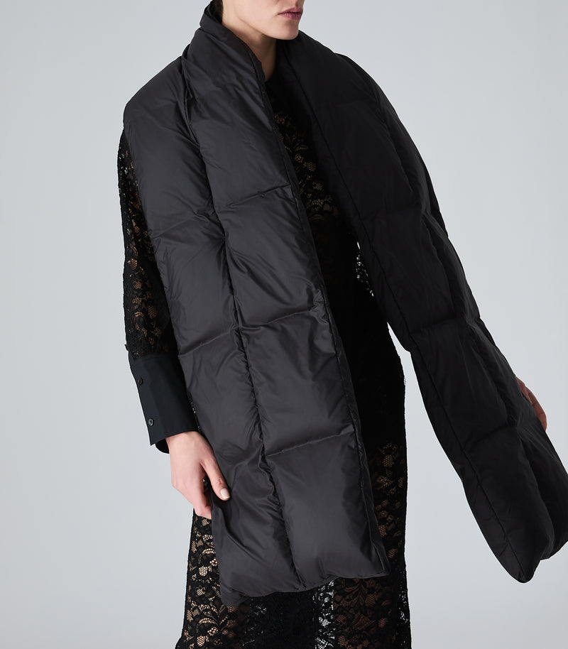 A model wearing a long black puffer vest over a black lace top, shown from the chin down against a light gray studio background.