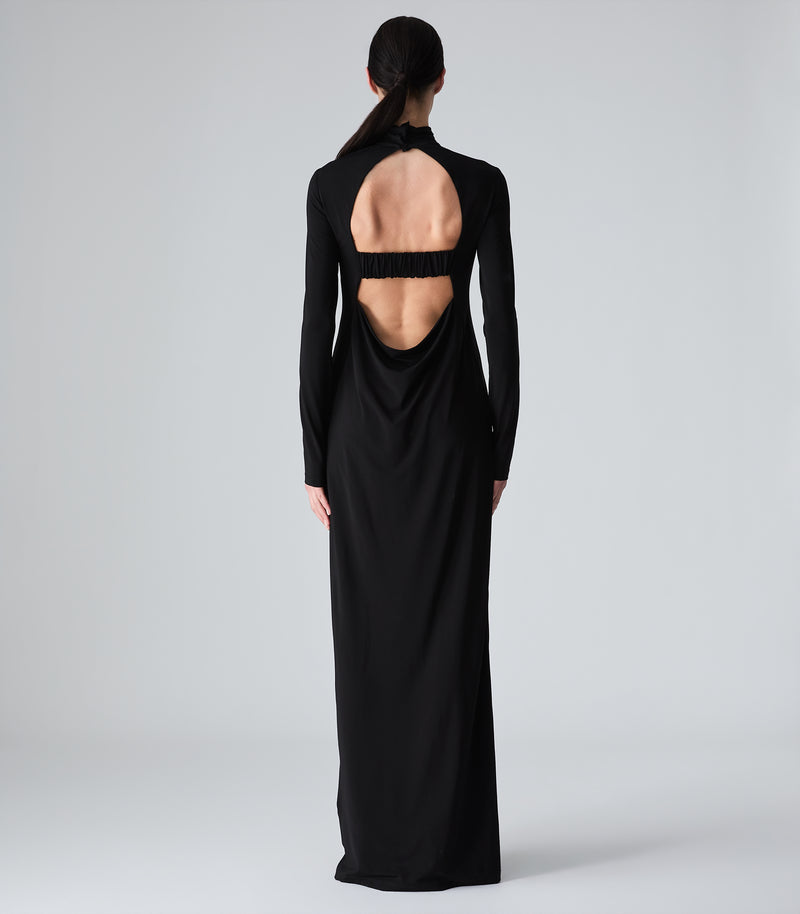 Full-length rear studio view of a model showcasing a black maxi dress to highlight its dramatic open-back design. The dress features a high, buttoned funnel neckline above a large oval cut-out that reveals the upper back, bisected by a horizontal, gathered elasticated modesty strap. The lower back drapes into a soft, fluid cowl shape before flowing into a long column skirt that hits the floor, set against a seamless light grey backdrop.
