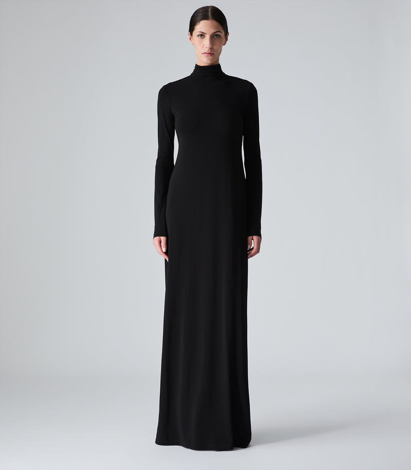 Full-length studio product view of a model presenting a minimalist floor-length black maxi dress. The design features a form-fitting column silhouette, a sophisticated high funnel neckline, and sleek, close-fitting long sleeves. The smooth matte fabric drapes fluidly to a straight hem that pools gently at the floor, captured against a seamless light grey backdrop.
