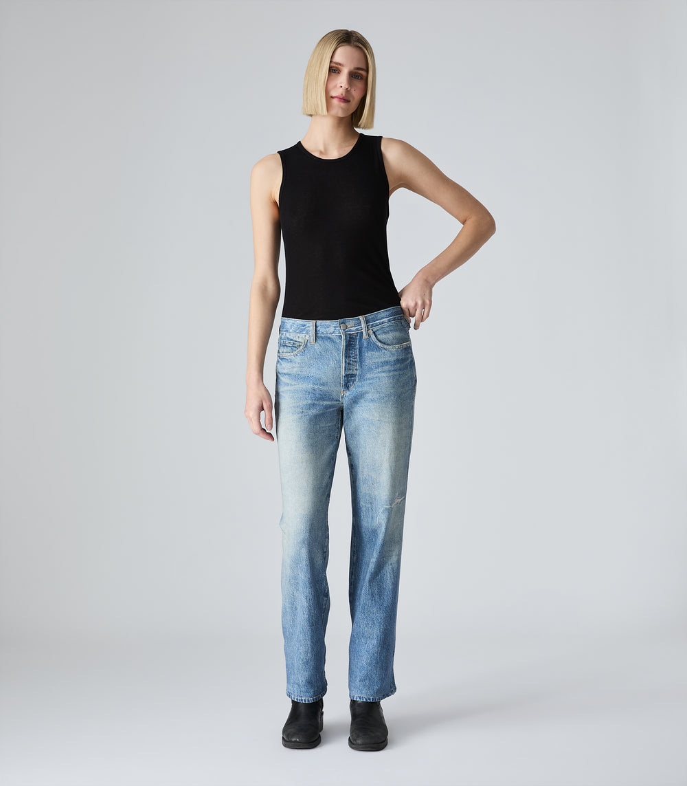 A woman modeling a black sleeveless ribbed tank top and light blue straight-leg denim-illusion jeans against a light gray studio background.