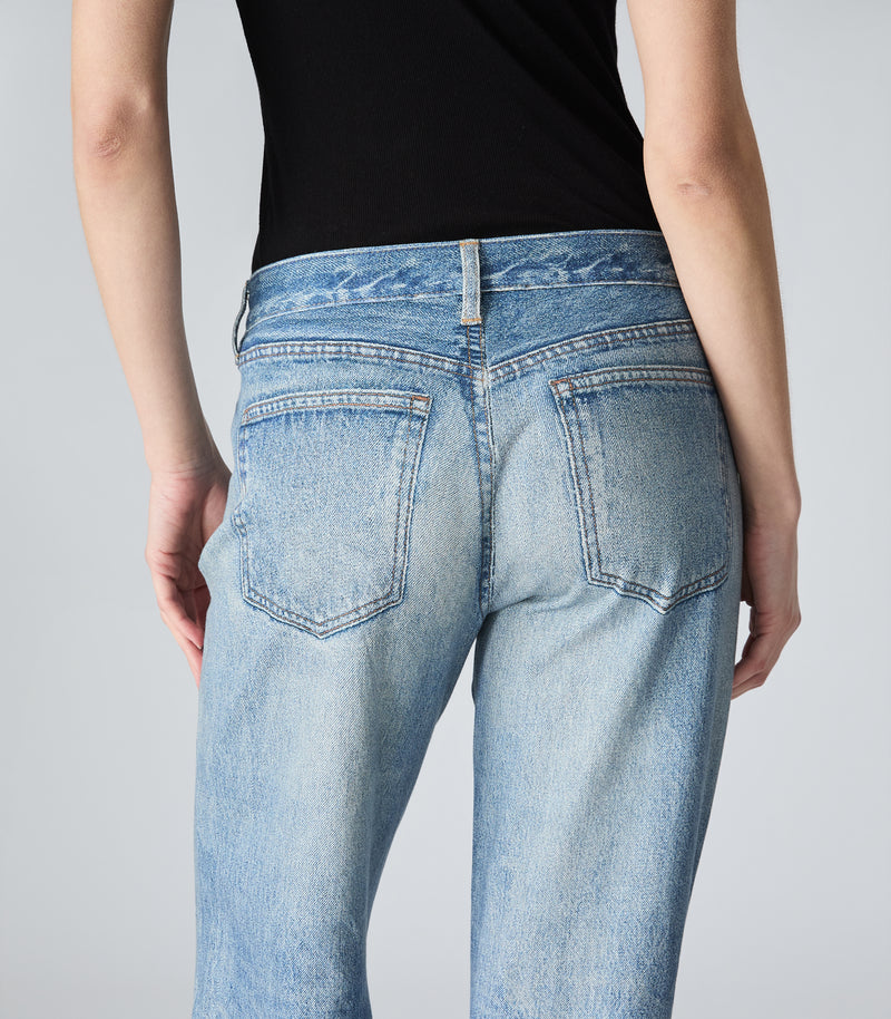 Rear view of a person wearing light-wash blue jeans with a black sleeveless top, shown against a plain light gray studio background.